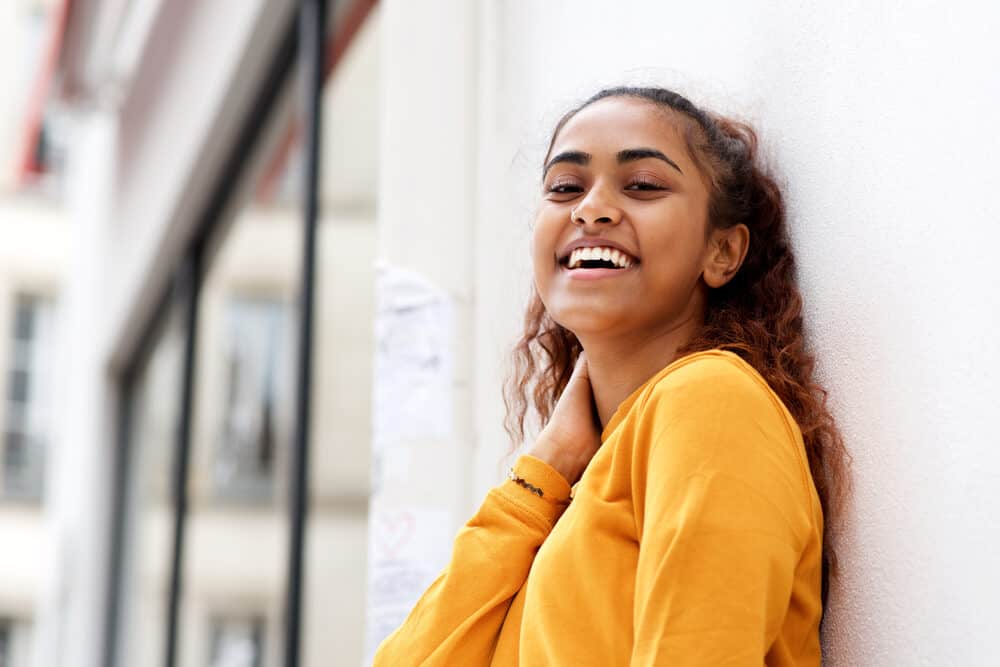 Vrouw van Indiaanse afkomst draagt een geelachtig oranje shirt en staat naast een witte muur