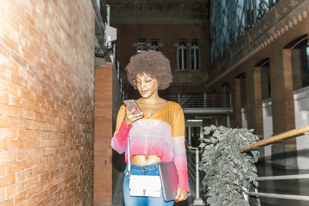 Een gelukkige Afro-Amerikaanse vrouw met donker haar in vrijetijdskleding controleert haar telefoon in een treinstation.