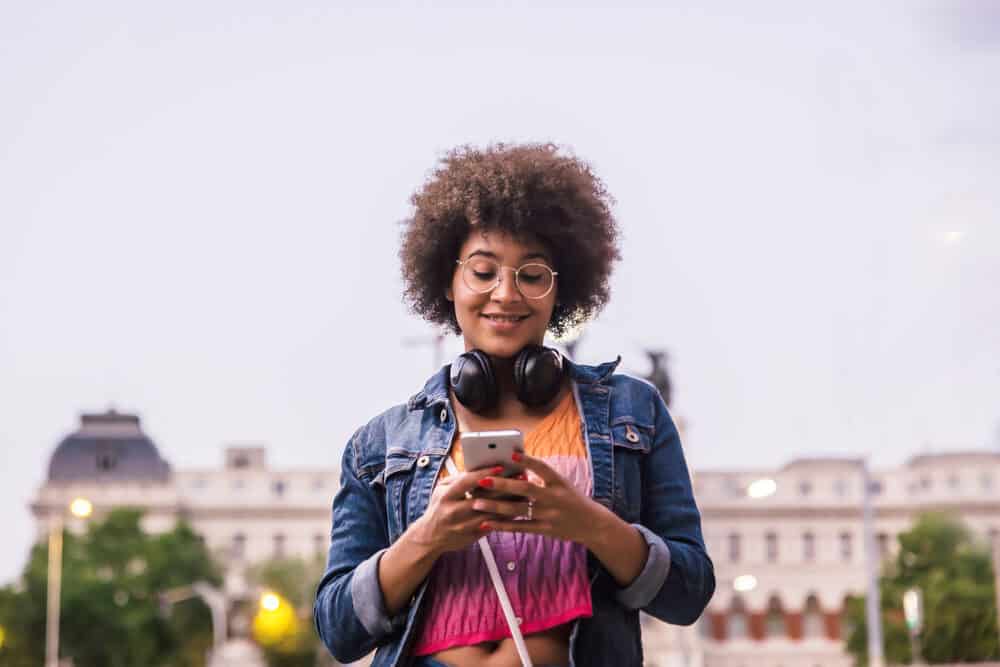 Een vrouw met een donkere huid en een zwarte koptelefoon lacht op straat naar haar smartphone in een shirt met levendige kleuren.