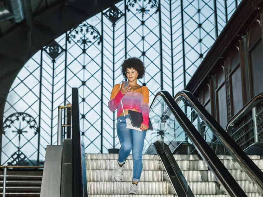 Een vrouw met donker haar kijkt op haar telefoon terwijl ze de trap afloopt in een treinstation.