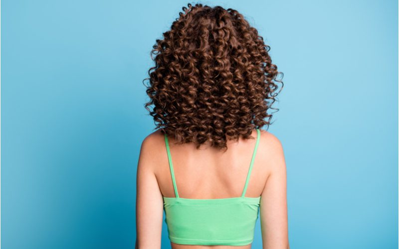 Afbeelding voor een stuk over hoe strakke krullen te krijgen met een vrouw in een blauwe kamer tegenover een muur met strakke krullen