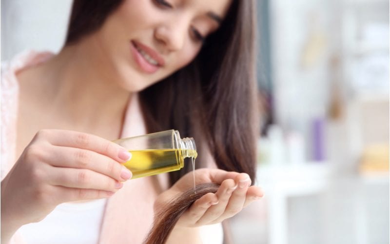 Voor een stukje over wat is haarserum, een brunette vrouw die wat van de olie in haar hand giet terwijl ze in een wit shirt staat met een wazige achtergrond