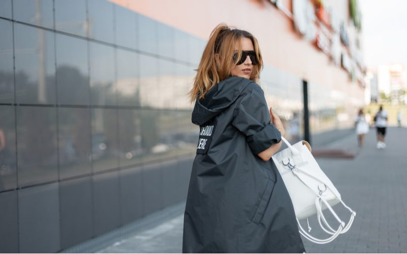 Vrouw in een lange trenchcoat loopt buiten bij een betegelde muur