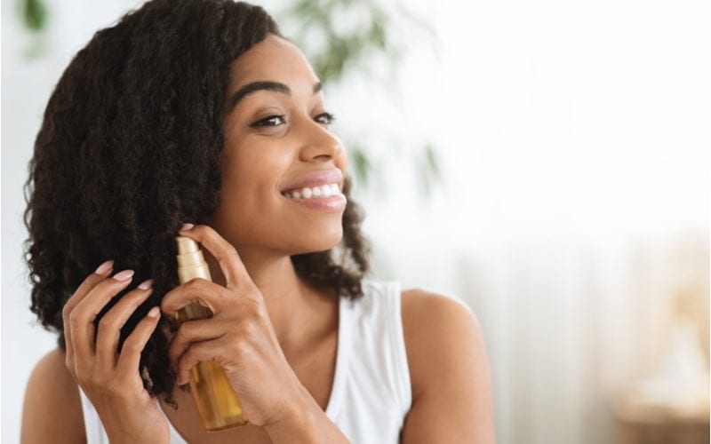 Gelukkige jonge Afro-Amerikaanse vrouw met natuurlijk haar die haarmelk op haar halflange haar spuit.