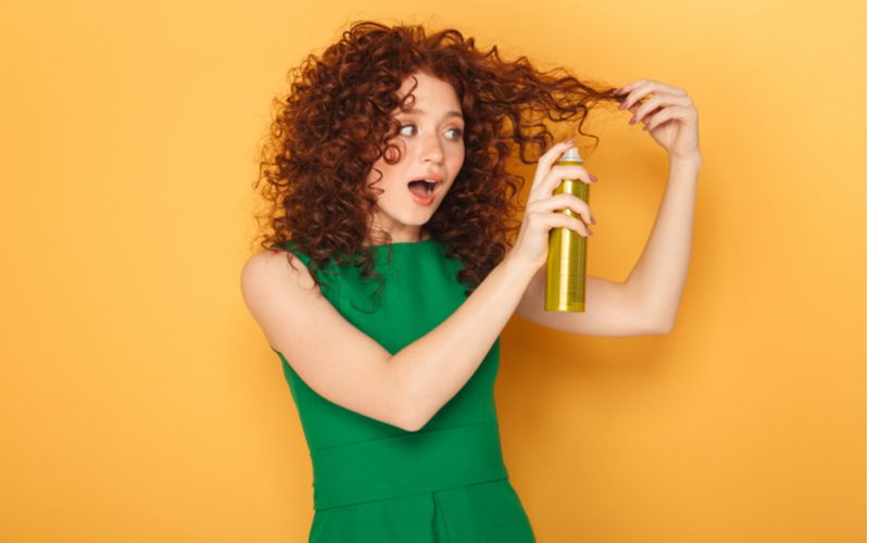 Vrouw spuit haar krullende rode haar met een haarverdikkingsproduct en staat in een groene jurk in een oranje kamer