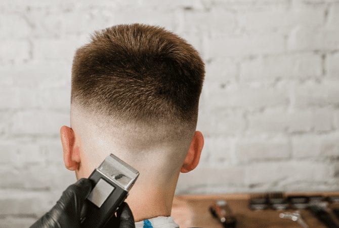 Man in een kapperstoel kijkt naar een bakstenen muur en krijgt een medium type fade kapsel van een gehandschoende kapper.