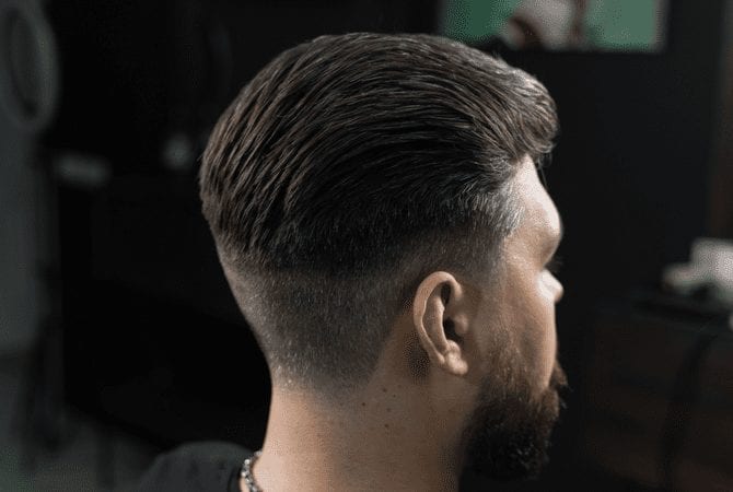 Een man met een lage fade en een slicked back top zit in een donkere kamer en kijkt weg van de camera.