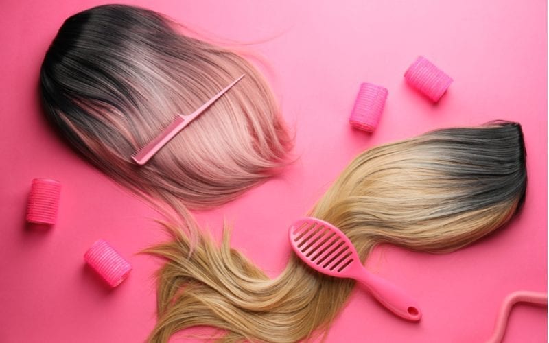 Voor een stuk over waar online goede pruiken te kopen, zitten twee haarstukken naast rollers en roze haartools bovenop een lay flat roze achtergrond.