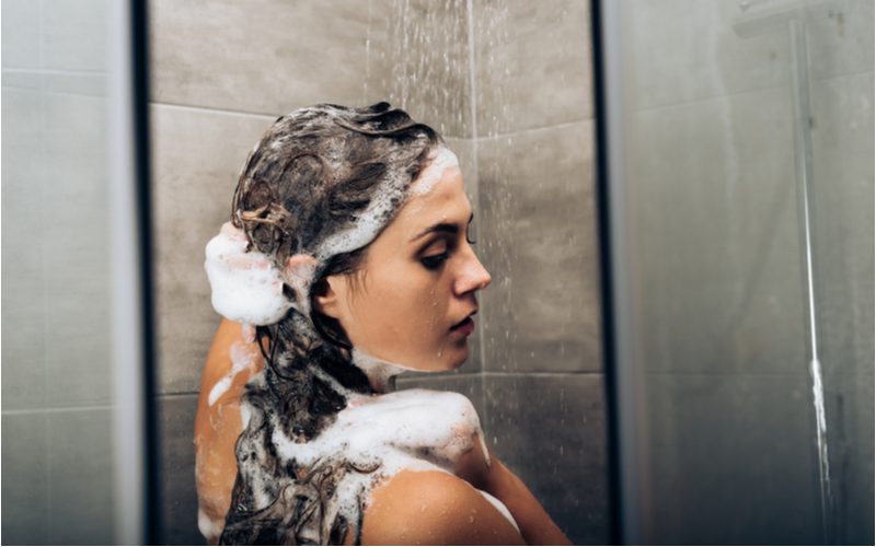 Meisje in een betegelde douche die haar lange haar inzeept terwijl ze over haar rechterschouder kijkt