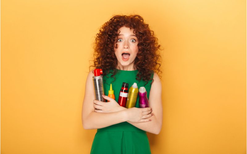 Portret van een vrouw met krullend haar in een groene jurk en een arm vol haarproducten in een oranje kamer.