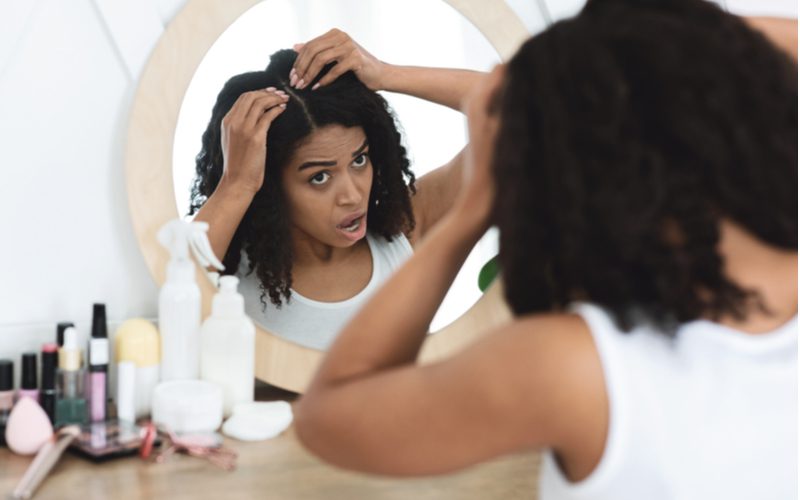 Vrouw op zoek naar haar hoofdhuid proberen te achterhalen waarom haar haar pijn doet terwijl staande het dragen van een witte tank top