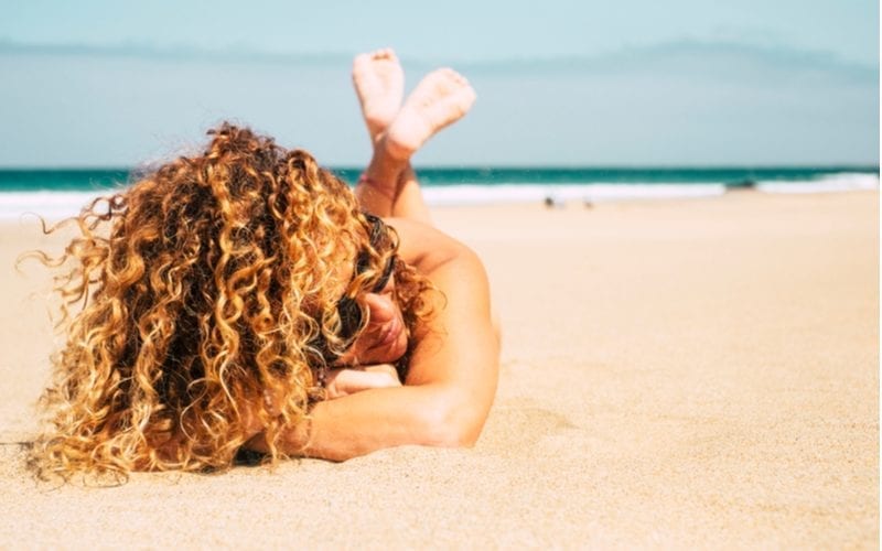 Een jonge vrouw liggend op haar buik op het strand met krullend haar dat ze in een vlecht moet doen omdat het lang genoeg is