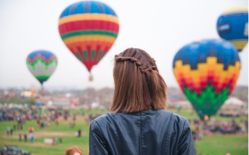 Vrouw met een kort lobbenkapsel staat weg van de camera en kijkt naar luchtballonnen met gevlochten kort haar