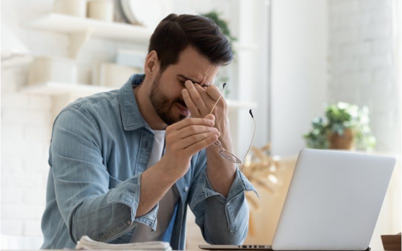 Man die gestrest is en zijn ogen vasthoudt met zijn bril in zijn andere hand met een open laptop als symbool voor een van de manieren om van roos af te komen (stress verminderen)