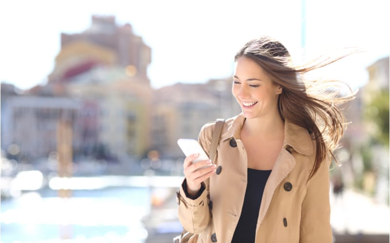 Gelukkig mooie vrouw lopen en schrijven of lezen sms berichten online op een smart phone terwijl de wind beweegt haar haar in een straat van een haven