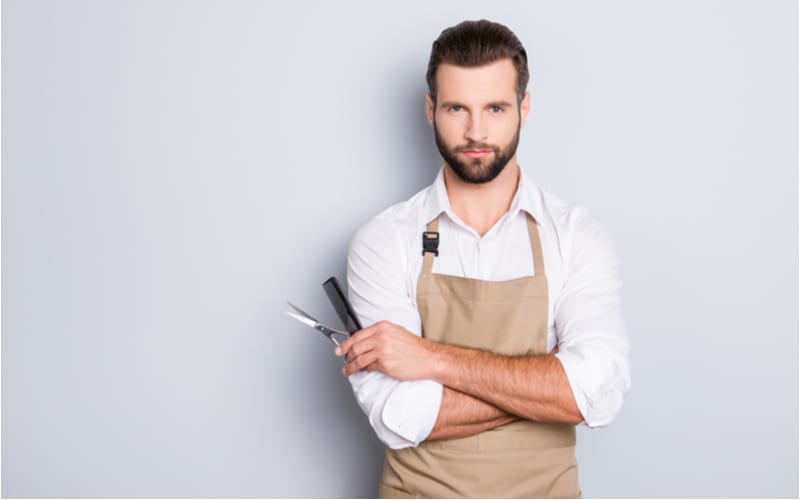 Portret met kopieerruimte, lege plaats van viriele harde kapper met zijn armen gekruist, apparatuur in de hand houdend, kijkend naar de camera, geïsoleerd op grijze achtergrond voor een stuk over wat een kapsalon is