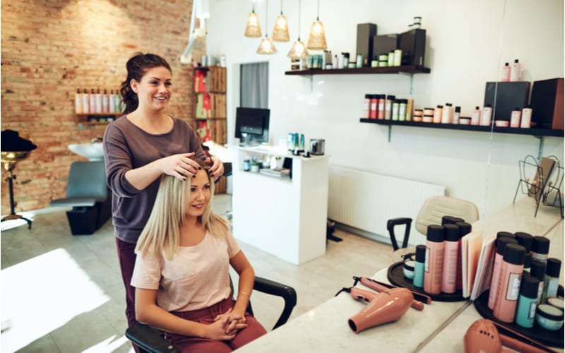 Jonge blonde vrouw die glimlacht en naar haar weerspiegeling in een spiegel kijkt terwijl ze in een salonstoel zit tijdens een afspraak met haar kapper