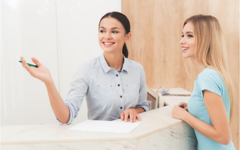 De donkerharige vrouw werkt bij de receptie. Ze werkt in een schoonheidssalon. Ze praat met de klant en laat iets zien.