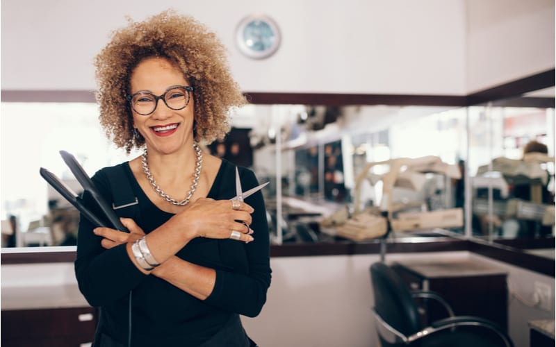 Professionele kapper die een stijltang en schaar vasthoudt. Vrouw kapper in gelukkige stemming in de salon als een stuk op het vinden van een kapsalon in de buurt van mij