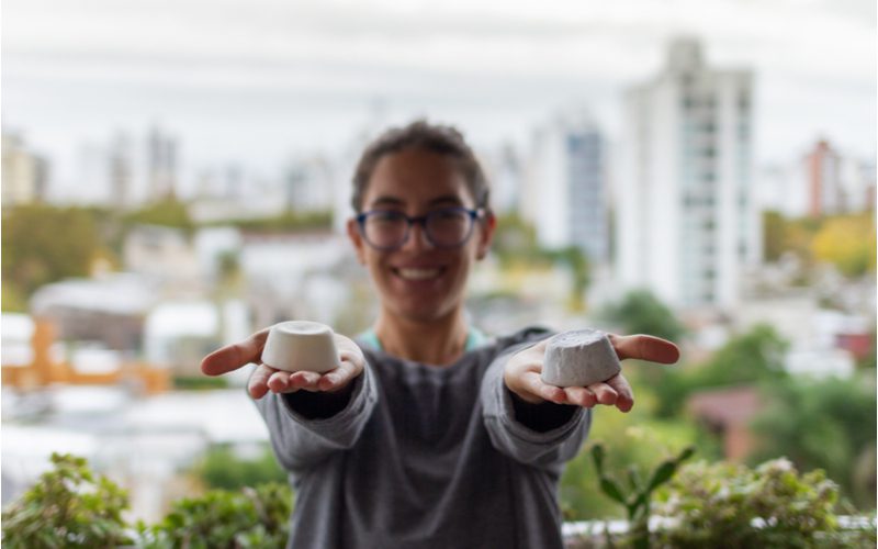 Mooie jonge vrouw die op een balkon staat te glimlachen en twee shampoo bars in beide handen houdt.