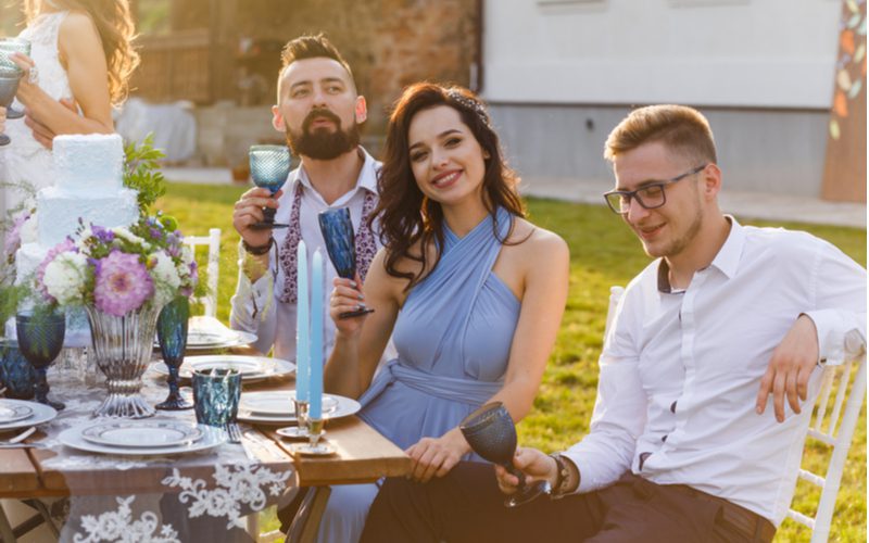 Afbeelding voor een stuk getiteld bruiloft gast kapsels met een man en vrouw zittend aan een rustieke tafel buiten om te proosten op de bruid en bruidegom.