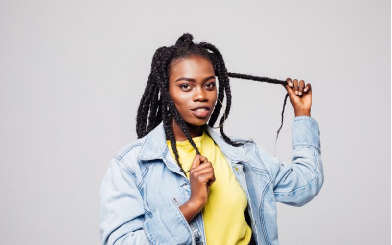 Voor een stuk over dreadlocks, een driestrengs gevlochten loc stijl op een vrouw die haar haar vasthoudt en een geel shirt draagt met een spijkerjasje.