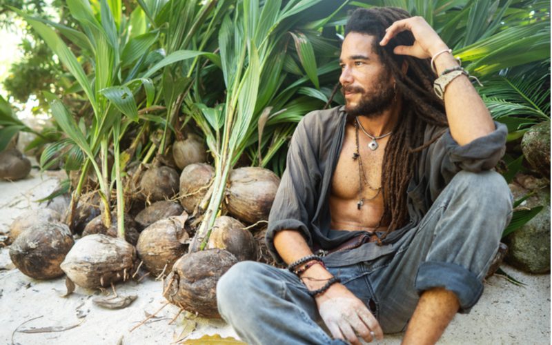 Island-aura man met wol gewreven dreadlocks hangend van zijn hoofd en zittend in kleermakerszit met zijn verweerde hemd open.