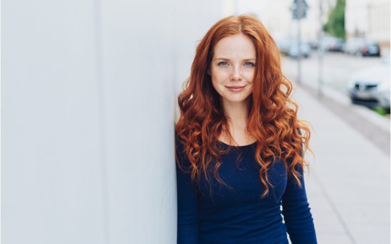 Bright Ginger-Toned Auburn haar op een dunne vrouw in een blauwe trui staande tegen een wit ommuurd gebouw.