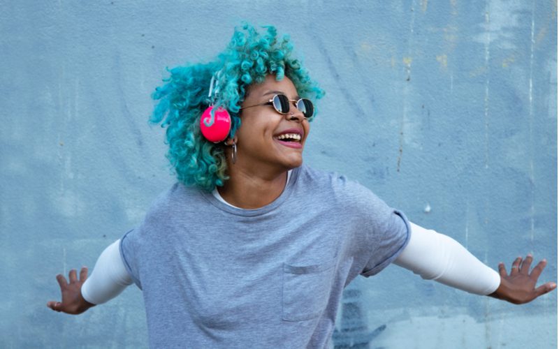 Aquamarijnblauwe haarkleur op een vrouw met bruine huid die een koptelefoon draagt en haar armen spreidt tegen een blauwe muur