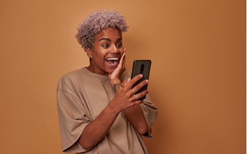 Violet-getint zilver haarkleur op een vrouw met bruine huidskleur die haar linkerwang in opwinding vasthoudt na het zien van iets wat ze leuk vond op haar smartphone.