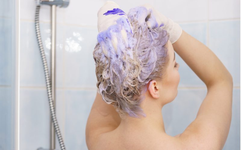 Bruinharige vrouw die paarse shampoo op haar haar gebruikt in een thuisdouche