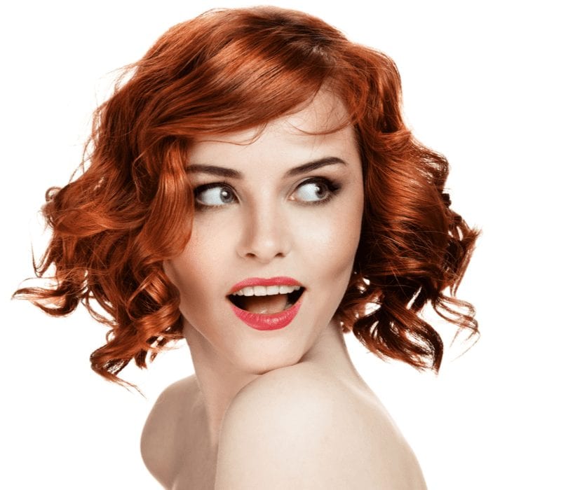 Dame glimlachend met rood haar en staande in een witte kamer tegen een witte muur en kijkend naar haar linkerzijde