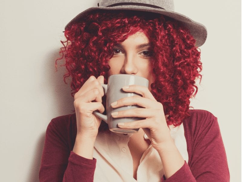 Dame met een boho hoed en rood haar dat gekruld is, staand en met een koffiekopje aan haar mond