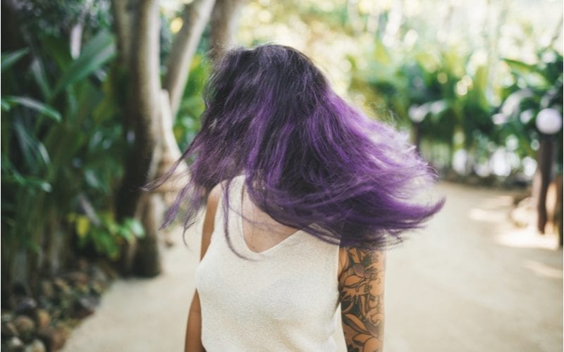 Vrouw met paars haar en een wit semi-transparant shirt met een tattoo mouw staat in een bos