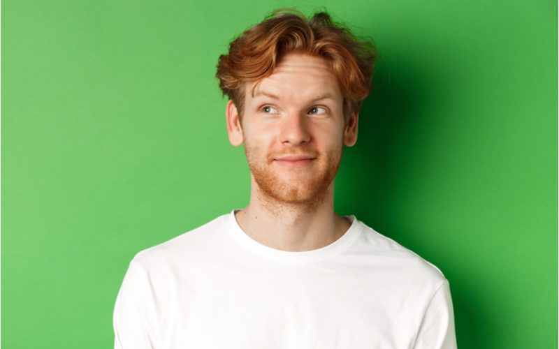 Split Wavy Mushroom Haircut op een man in een wit shirt met een grijns op zijn gezicht terwijl hij omhoog en naar rechts kijkt in een groene kamer.