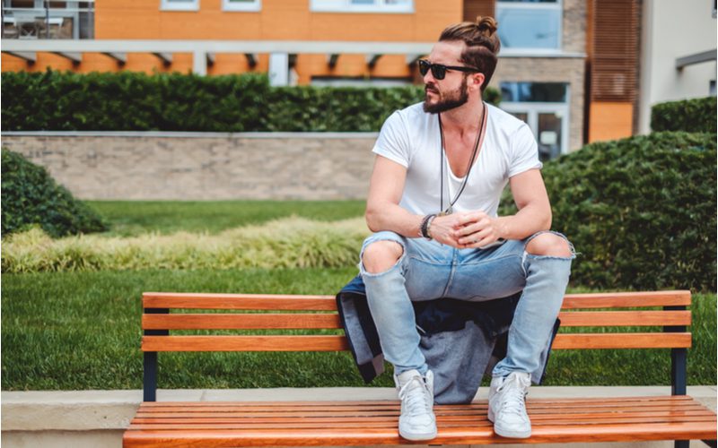 Man met een man bun en een dikke baard zit op de rug van een bank en kijkt naar rechts terwijl hij zijn handen in elkaar klemt.