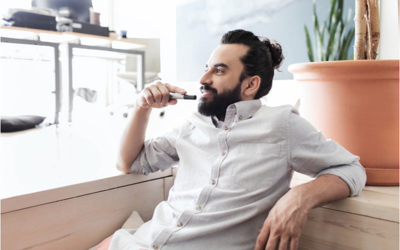 High-Volume Boosted Man Bun gedragen door een man in een witte button-up leunend tegen de achterkant van een gebruinde bank.