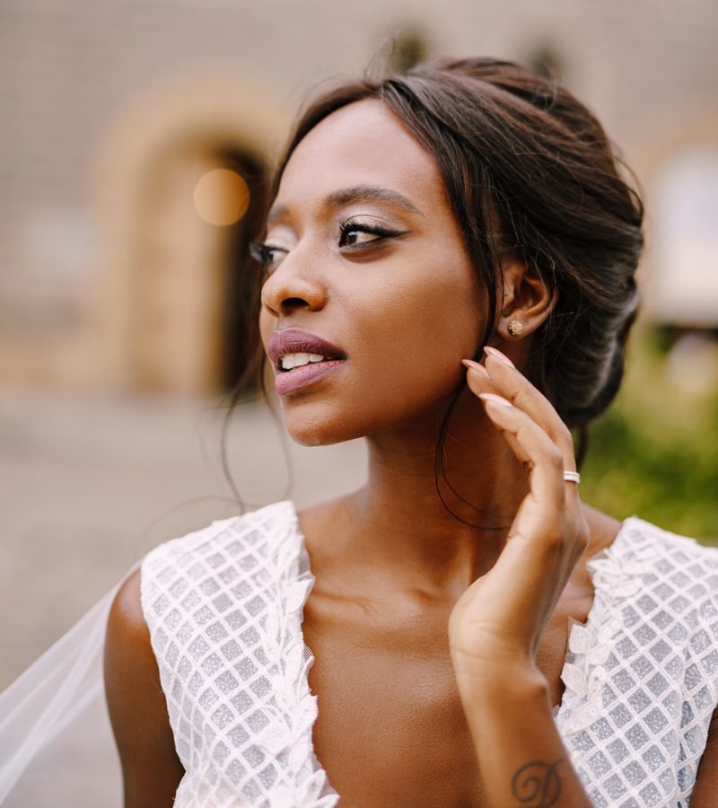 Foto voor een roundup op de beste Afro-Amerikaanse Bruidskapsels met een vrouw die op een rustiek oud stadsplein staat en haar hand tegen haar oor houdt.