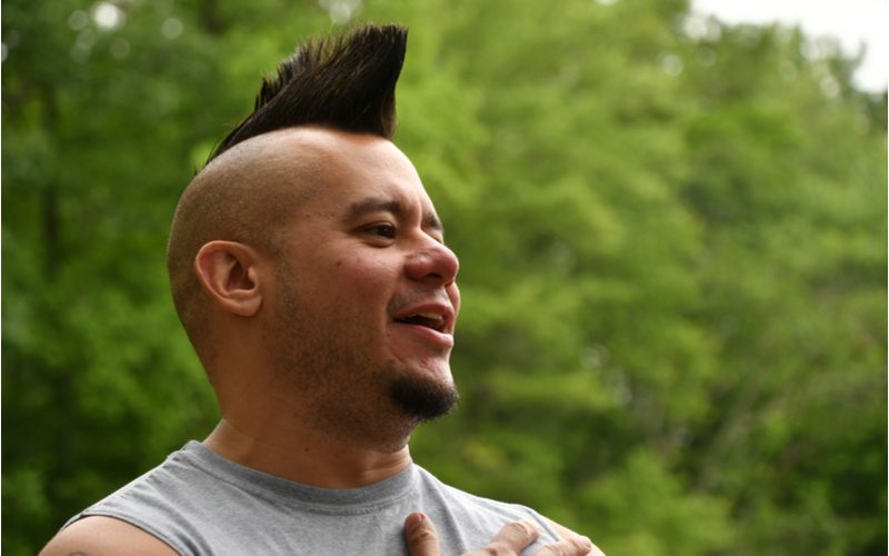 Psychobilly Wedge Mohawk Kapsel op een man in een afgeknipt grijs tshirt in een veld met bomen.