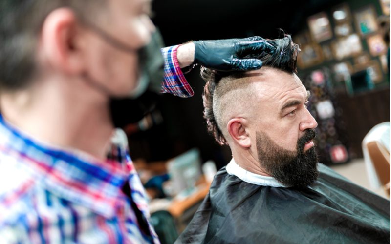 Man in een kappersstoel krijgt een mohawk kapsel van een gemaskerde kapper met handschoenen en een blauw en rood geruit hemd.