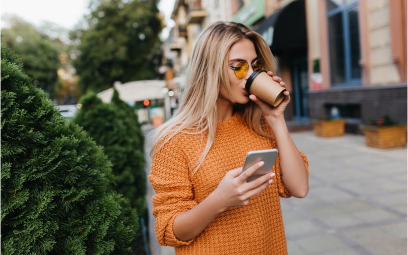 Face-Framing Fringe pony gedragen door een vrouw in een oranje trui die haar telefoon controleert en koffie drinkt.