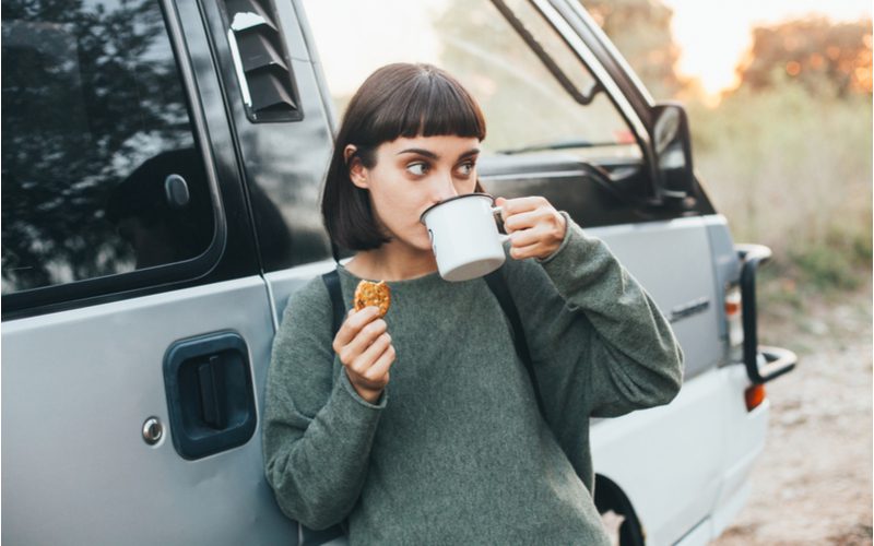 Een vrouw met een donkergroen shirt drinkt koffie uit een mok en leunt tegen een camper terwijl ze micro bangs draagt, één van de meest populaire soorten bangs.