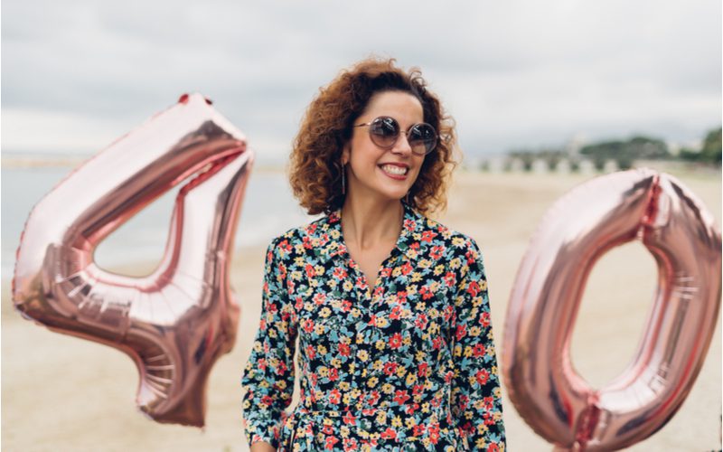 Voor een stuk over de beste kapsels voor vrouwen boven de 40, staat een meisje met een bril op het strand tussen ballonnen met de tekst 40