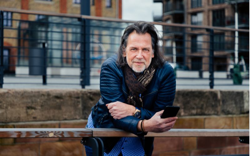 Oudere man met lang haar leunt over een metalen reling en kijkt naar zijn telefoon