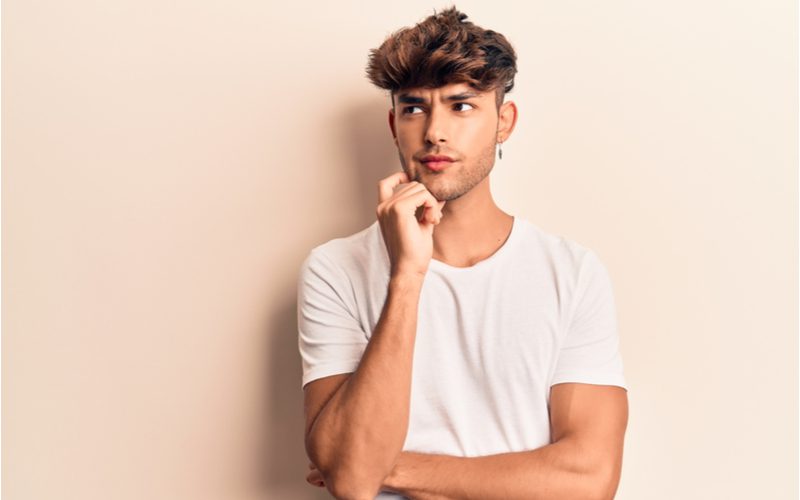 Feathered Two Block Haircut (aka Tiktok haar) op een man in een bruine kamer in een wit shirt die zijn rechterwang vasthoudt.