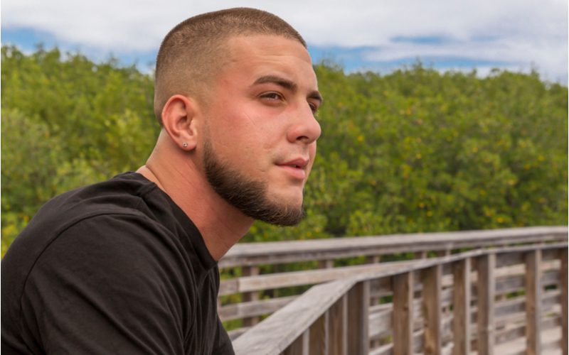 #2 Bald Drop Fade, een van de meest populaire kapsels voor mannen, op een man die buiten staat op een houten promenade in een natuurgebied.