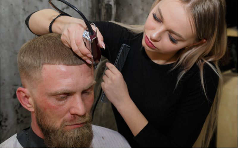 Man in een kappersstoel krijgt een Lined-Up Low Bald Caesar Fade kapsel van een vrouwelijke kapper.