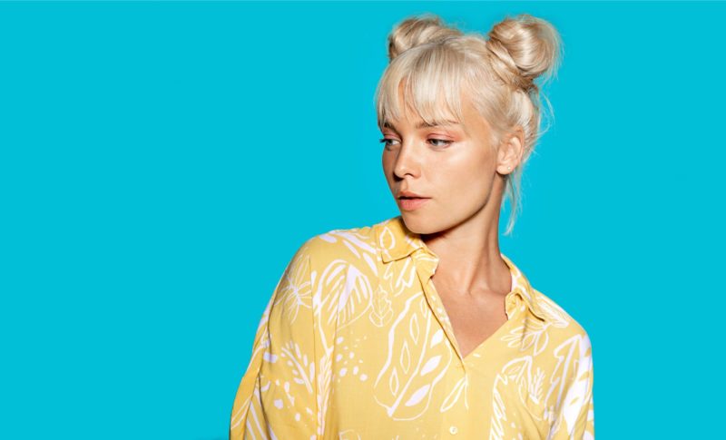 Blonde vrouw in een blauwe kamer en een geel shirt met volwassen space buns, een kenmerkend lang tot halflang kapsel.