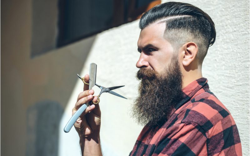 Long Comb Over Skin Fade With Hard Part op een houthakker uitziende man die een baard trimschaar vasthoudt.