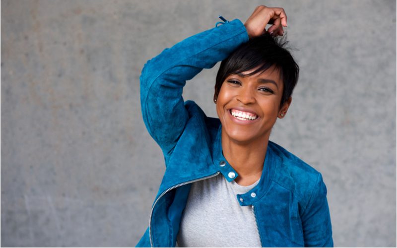 Pixie Black Bob Haircut With Side-Swept Bangs gedragen door een vrouw in een blauw suède jasje die haar haar op de top houdt.